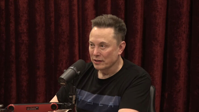 Elon Musk, one of the rudest celebs, in a dark t-shirt speaking into a microphone with a red curtain background.