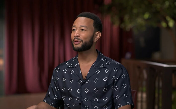 John Legend, a rude celeb, in a dark patterned shirt, speaking during an interview against a blurred red curtain background.