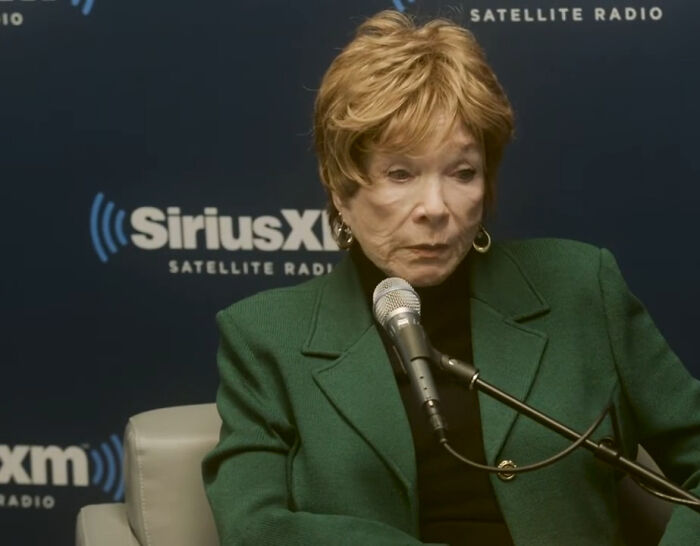 A celeb with light brown hair, wearing a green jacket and black turtleneck, speaking into a microphone; some rudest celebs.