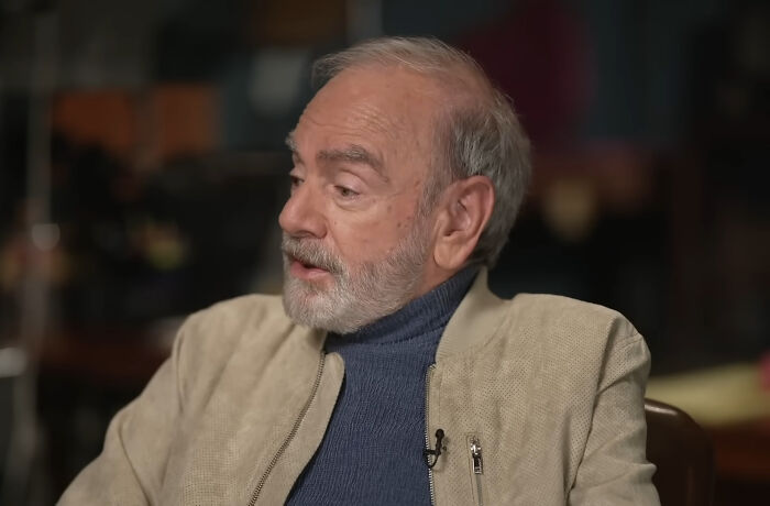 A grey-haired man with a beard wearing a beige jacket and a blue turtleneck, possibly a rude celeb.