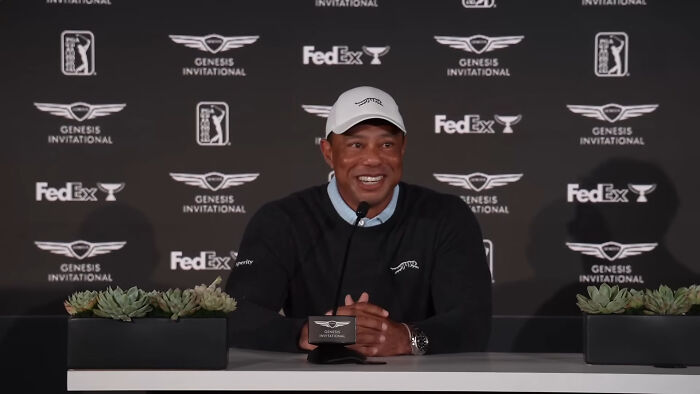 Tiger Woods, wearing a white cap and black sweater, smiles while speaking at an event, perhaps a rude celeb.