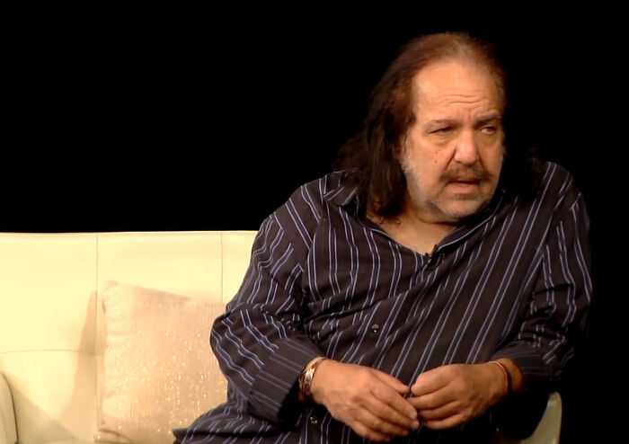 A man with long dark hair and a mustache, wearing a striped shirt, sitting on a white couch. Rudest celebs.