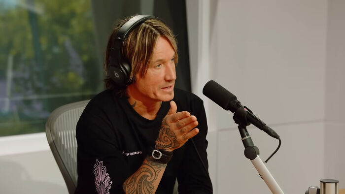 Country music celeb Keith Urban, wearing headphones and a black shirt, gesturing while speaking into a microphone.