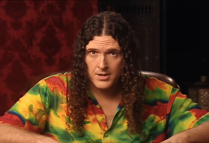 Al Yankovic, a celebrity with long curly hair and a tie-dye shirt, looks directly forward, perhaps one of the rudest celebs.