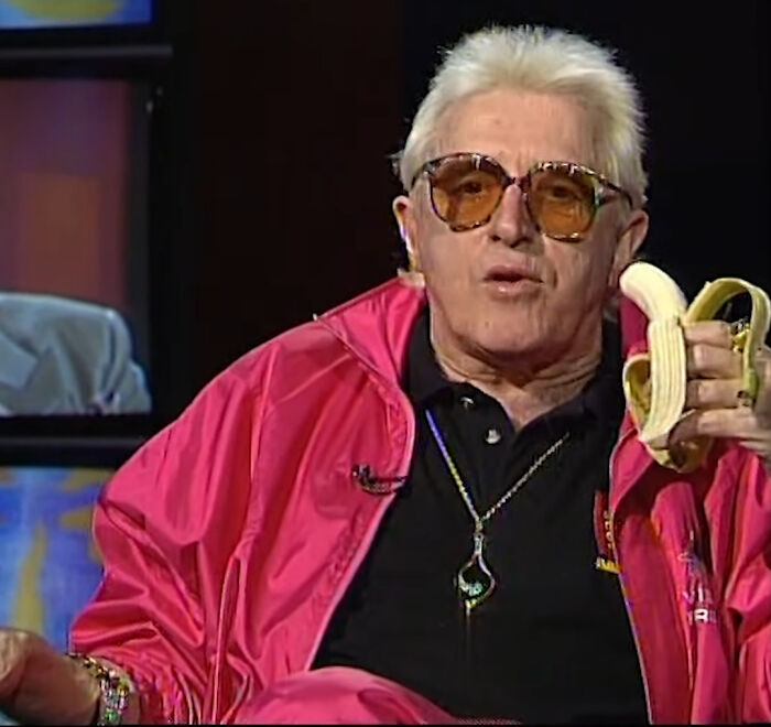 A man with white hair, amber glasses, and a pink jacket holds a peeled banana. He could be one of the rudest celebs.