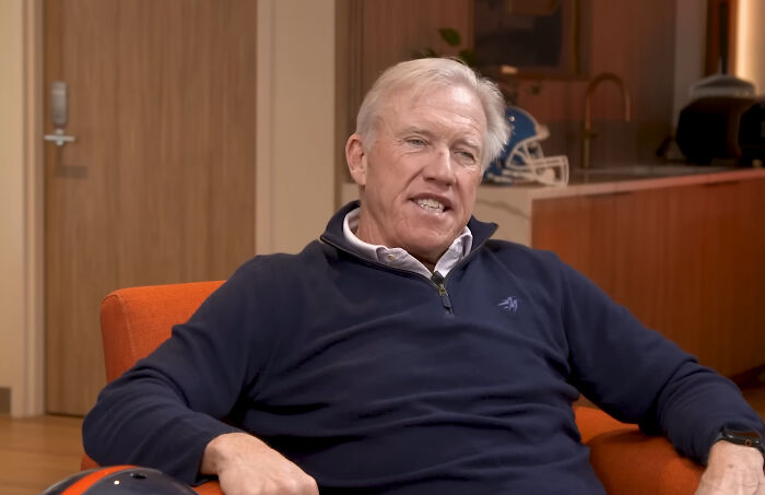 An older white man with white hair, wearing a navy blue quarter-zip sweater, sits on an orange chair. He could be one of the rudest celebs.
