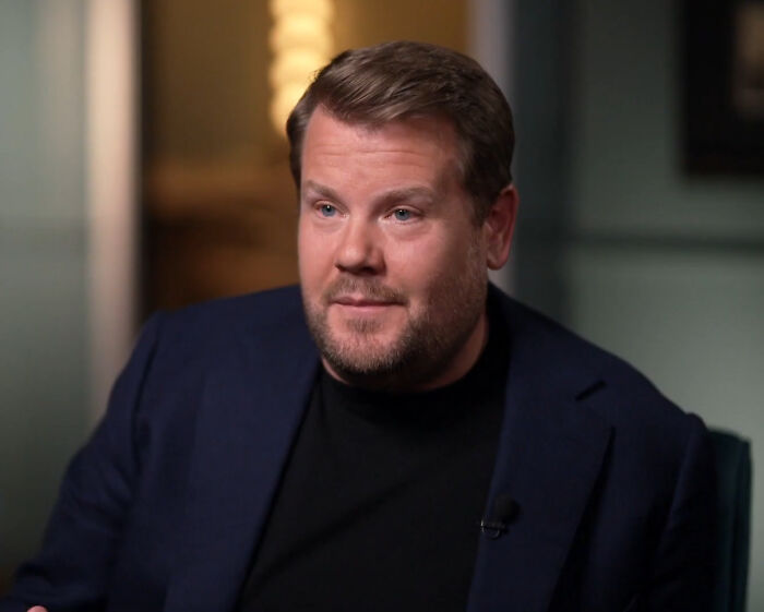 James Corden, a celebrity often called rude, with a beard and dark blue suit jacket. He looks serious while sitting.