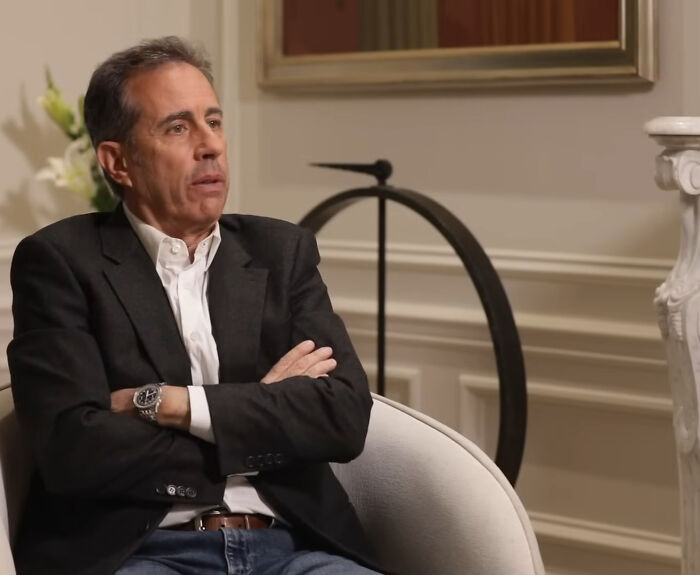 Jerry Seinfeld, one of the famous celebs, sitting with arms crossed, wearing a dark jacket, white shirt, and jeans.