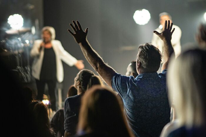 Audience members raising hands in a dimly lit event, capturing a moment of normal family activities that seem bizarre now.
