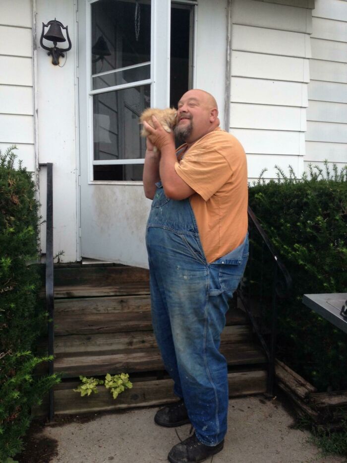 A smiling man in dirty overalls holding a tiny kitten to his face, reflecting farm life that won't make sense to city kids.
