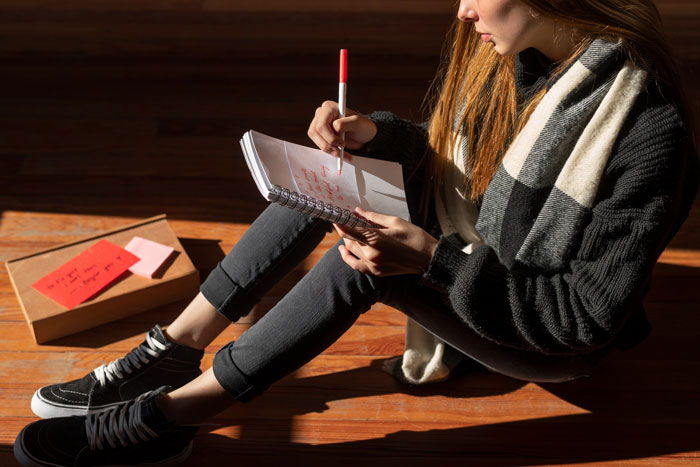 Young woman sitting on floor writing in notebook with pen, involved in prisoner pen pal letter on behalf of rude neighbor.