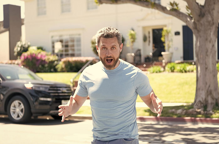 Man in a blue t-shirt angrily confronting a neighbor outside a suburban home on a sunny day, showing frustration.