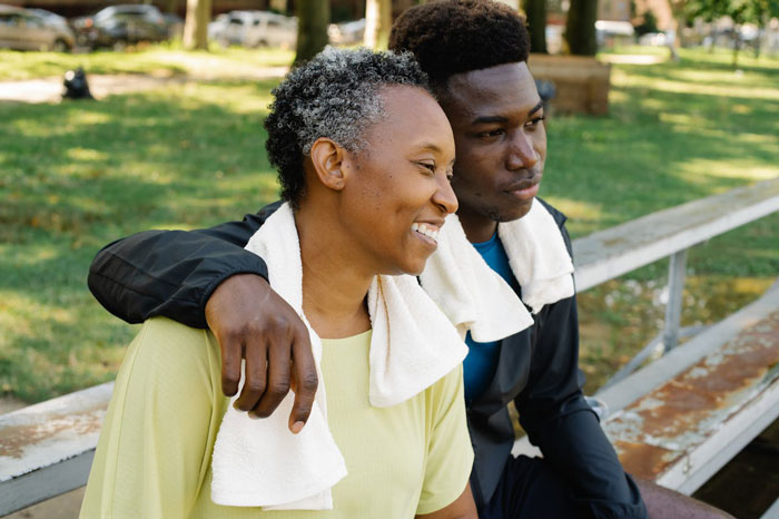 Couple sitting on a bench outdoors with towels around their necks after exercising, showing warmth and support.