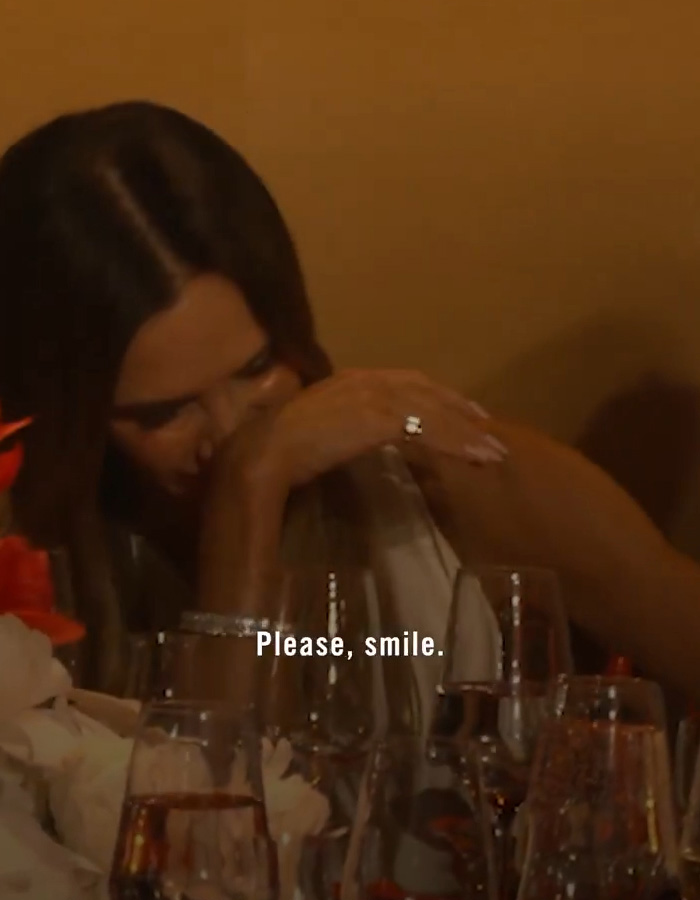 O rosto sério de Victoria Beckham, parcialmente obscurecido, com texto Por favor, sorria. Uma abordagem humorística de sua expressão característica.