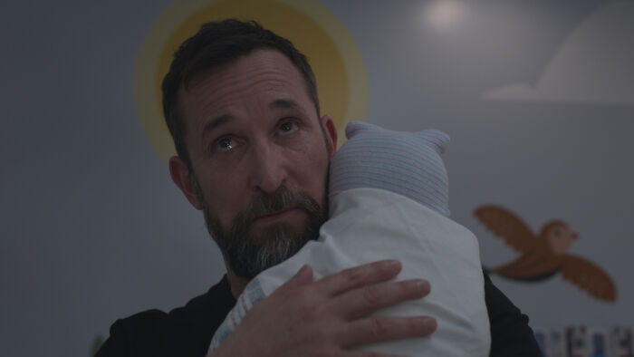 Man with beard holding a baby wrapped in a blanket, scene from The Pitt Season 3 showing emotional moment indoors