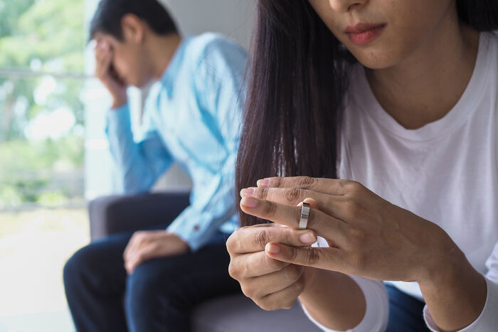 Woman removing wedding ring looking upset while man sits in background holding his head, reflecting on childhood norms.