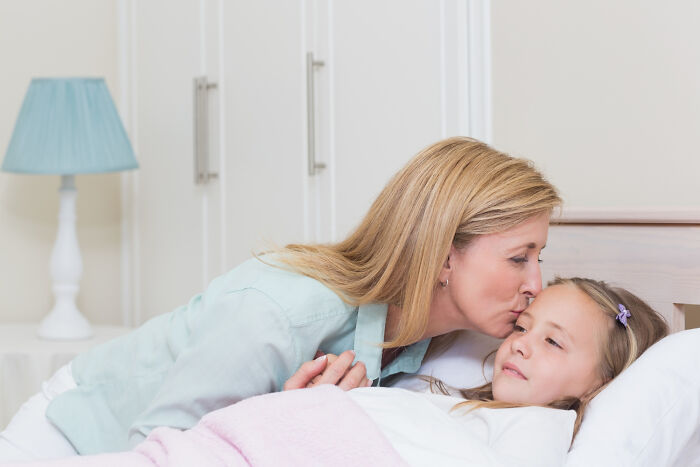 Mother kissing her daughter goodnight in bed, illustrating nostalgic childhood norms people later found weird.