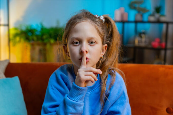 Young girl sitting on a couch with finger to lips, representing childhood norms that seem strange in hindsight.
