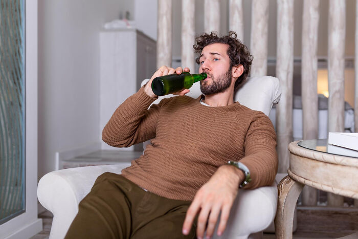 Young man relaxing on a white armchair drinking from a green bottle reflecting on childhood norms now seen as weird.