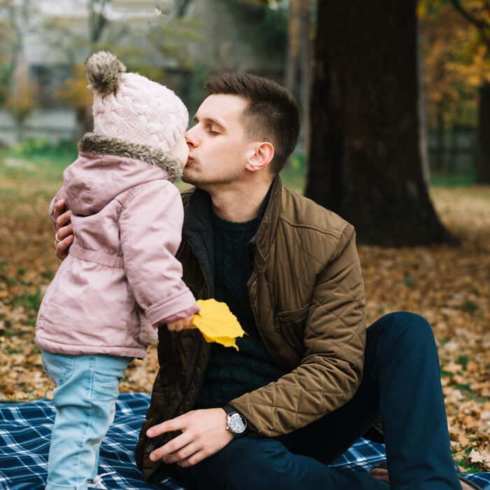 Man sitting on a blanket giving a kiss to a toddler wearing a pink jacket and hat during a fall outdoor moment childhood norms.