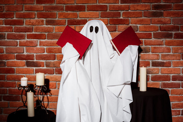 Person dressed as a ghost holding two red books, surrounded by lit candles against a brick wall, childhood norms concept.
