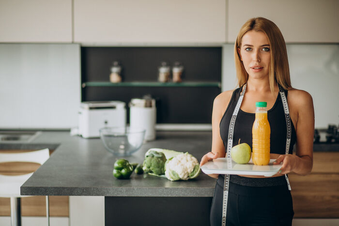 Woman in athletic wear holding a plate with apple and juice, surrounded by vegetables, highlighting childhood norms and healthy habits.