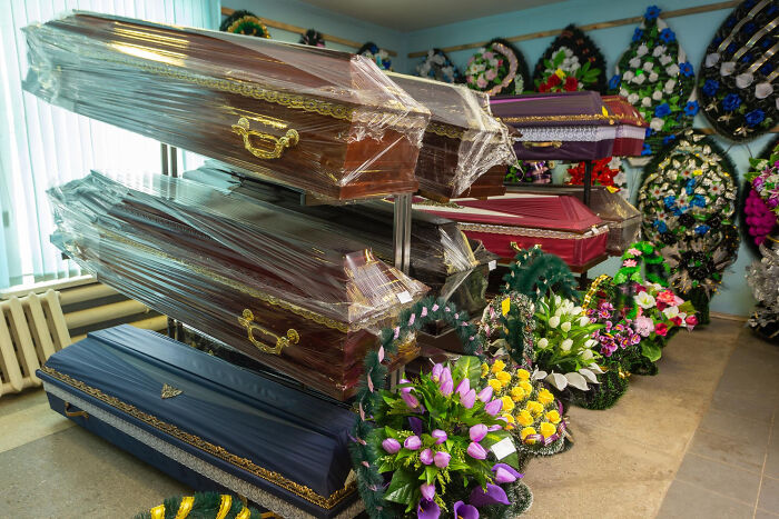 Stacked coffins wrapped in plastic with colorful flower wreaths displayed in a funeral home setting, showing childhood norms weirdness.