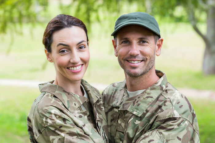 Smiling man and woman in camouflage uniforms outdoors representing childhood norms through military family life.