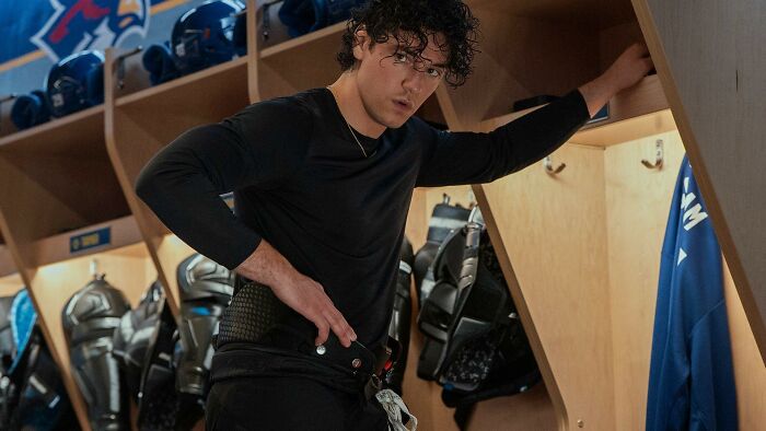 A man in a locker room, wearing a black shirt and athletic gear, looks intently at the camera. Steamy Romance.