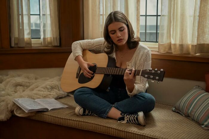 A young woman playing an acoustic guitar, cross-legged on a bench, for Prime Video's new romance.