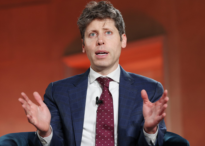 OpenAI CEO Sam Altman speaking during an event, wearing a blue suit and red polka dot tie with hand gestures.