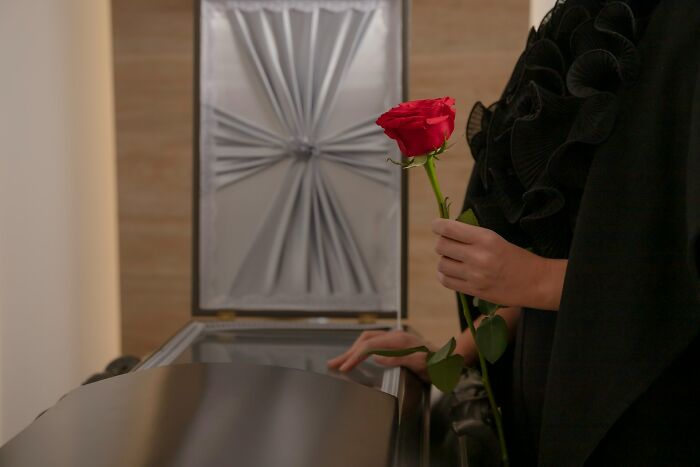 A person in black holds a red rose by an open coffin. Perhaps a MIL passed away.