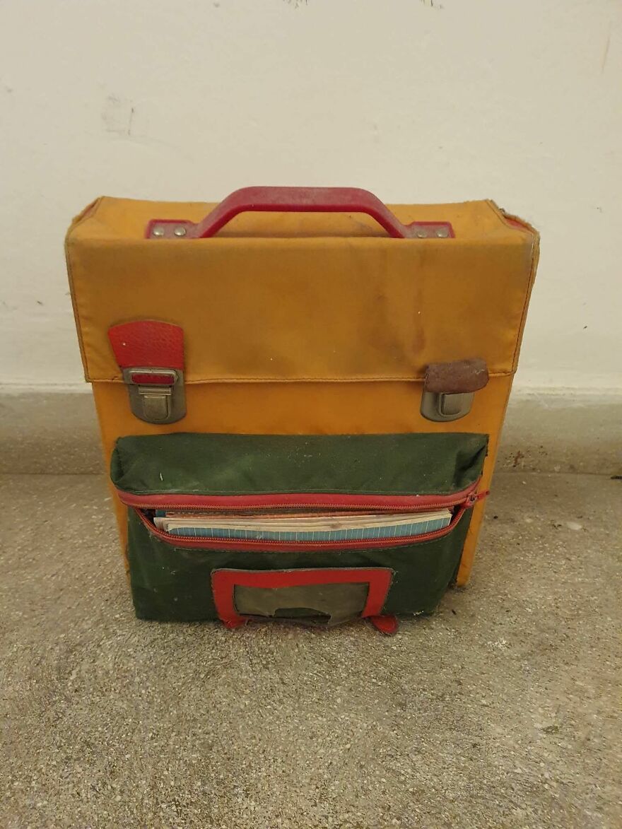 Old yellow and green bag with red handle and latches, showing pareidolia as it resembles a face, on a concrete floor.
