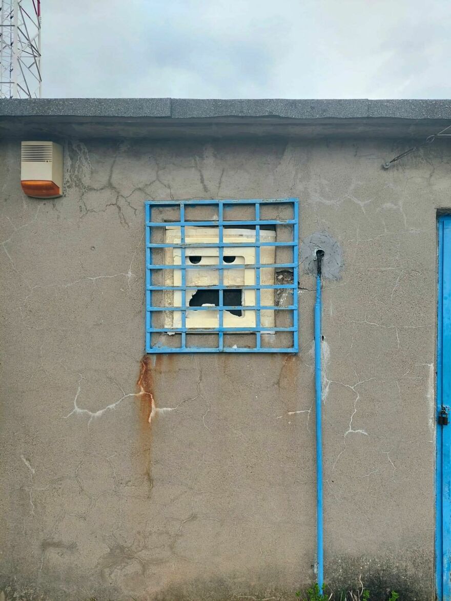 Rusty barred window and cracked wall creating a face pareidolia effect on a grey building exterior.