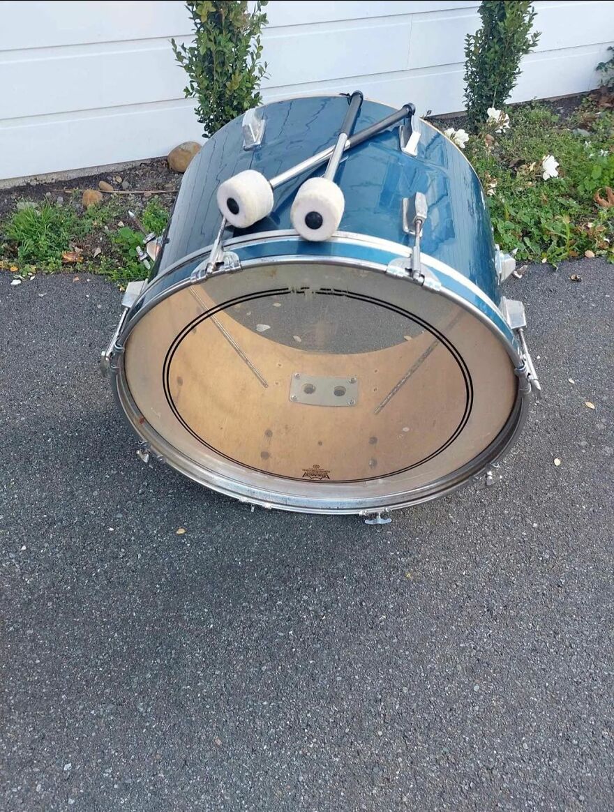 Bass drum and mallets resembling eyes, creating a pareidolia effect on a paved surface near plants outdoors.