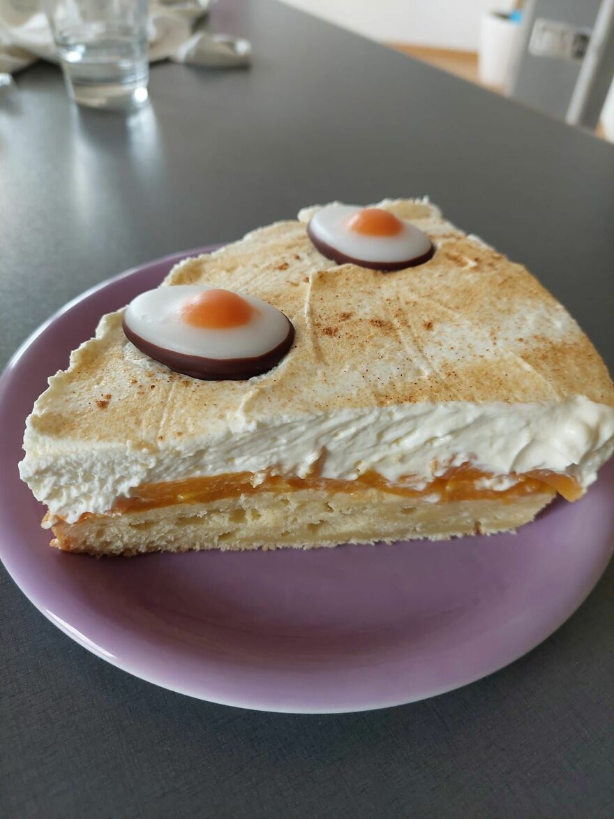 Slice of dessert with candy shaped like fried eggs creating a pareidolia effect of a face on a purple plate.