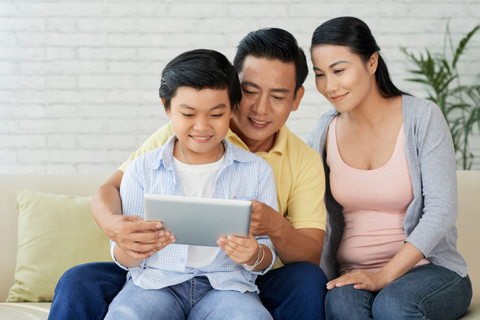 Asian couple encourages their son to be independent at 18, sitting together and smiling as he uses a tablet on the couch.