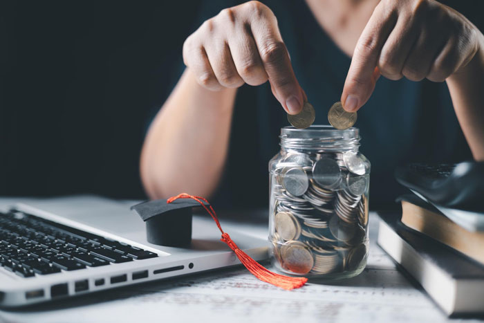 Person placing coins in jar filled with change near laptop and graduation cap symbolizing independence and financial responsibility.