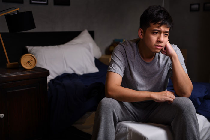 Young man sitting on bed looking thoughtful and upset, reflecting on being pushed to be independent at 18 years old.