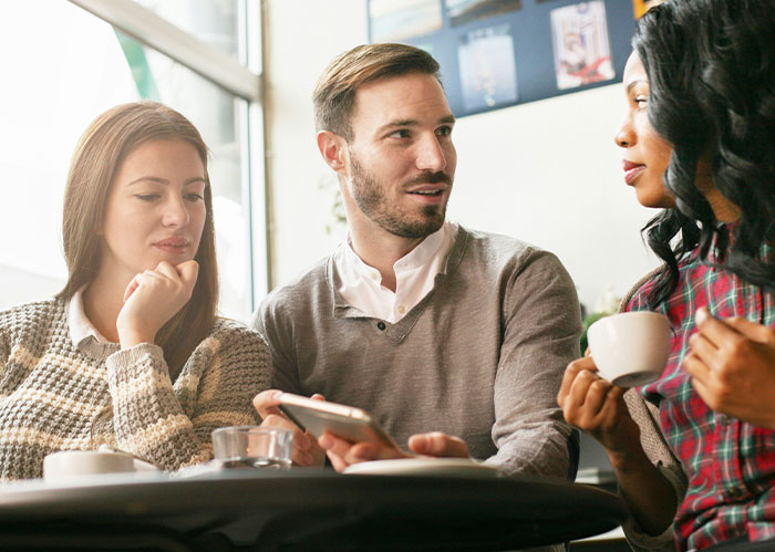 Three friends discussing, one looking at a phone, one holding a coffee, contemplating needing better friends.