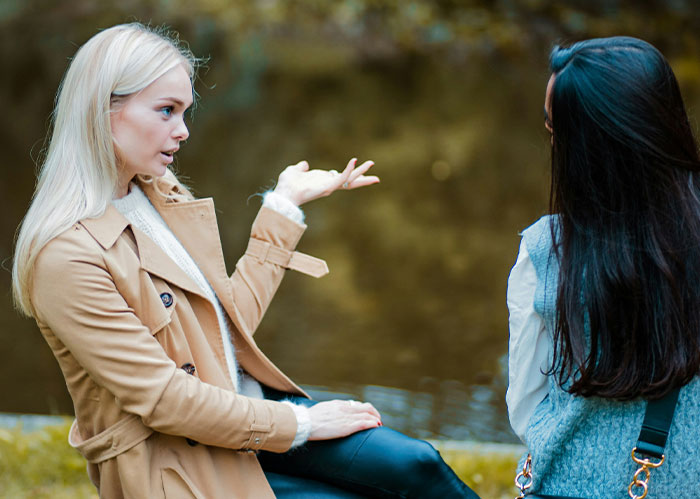 Blonde woman in trench coat gesturing while talking to a friend, reflecting on needing better friends.