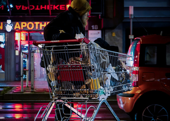 A person with a beard and hooded jacket pushing a shopping cart full of belongings at night, needing better friends.