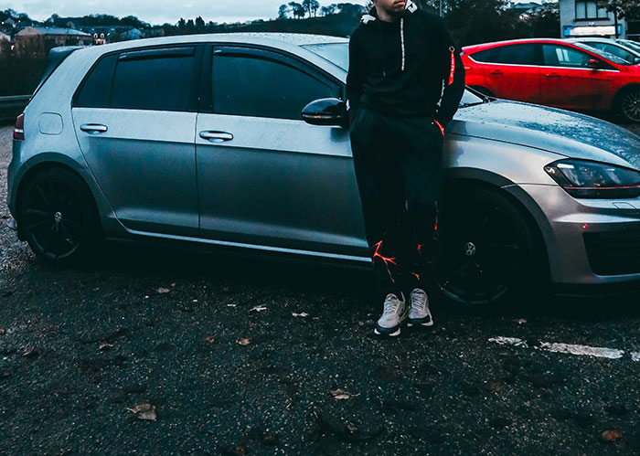 A man in black athleisure with reflective accents leans on a silver car in a parking lot, needing better friends.