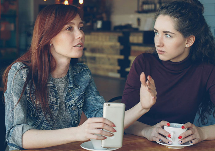 Two women in a cafe. One, in a denim jacket, speaks while the other, with curly hair, gestures, realizing she needs better friends.