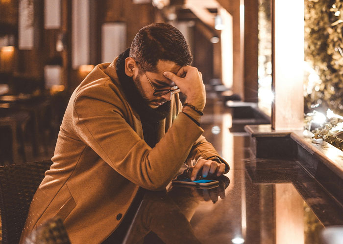 A man in a tan coat and glasses, looking distressed while checking his phone. He needs better friends.