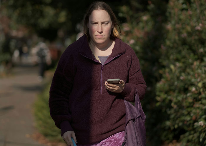 A woman walks outdoors in a burgundy fleece, looking at her phone, realizing she needed better friends.