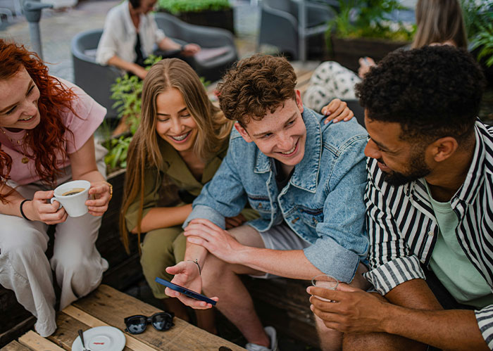 Four friends laughing and looking at a phone, one holding coffee. They realize they need better friends.