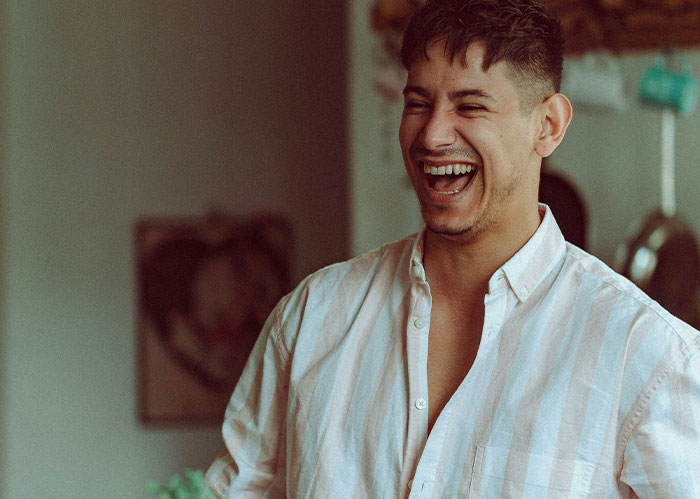 A man with short dark hair and a striped shirt laughs heartily, open-mouthed, needing better friends.