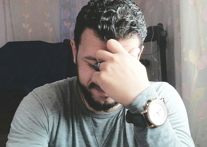 A man with dark curly hair and a beard, wearing a grey shirt and watch, looking distressed with his hand on his face. He needs better friends.