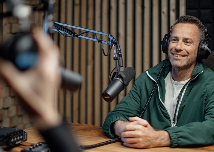 A smiling man with headphones and a green jacket records a podcast. He realized he needed better friends.